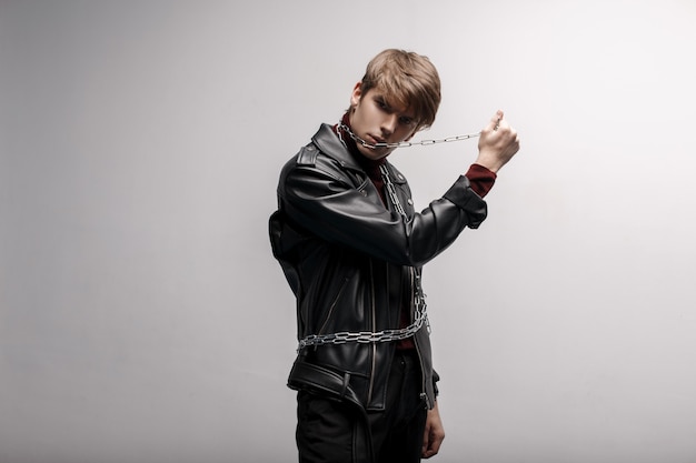 A young hipster man with a fashionable hairstyle in red golf in a black stylish jacket in black jeans holds a metal chain in his hands and poses for the camera near a white wall. Fashionable guy