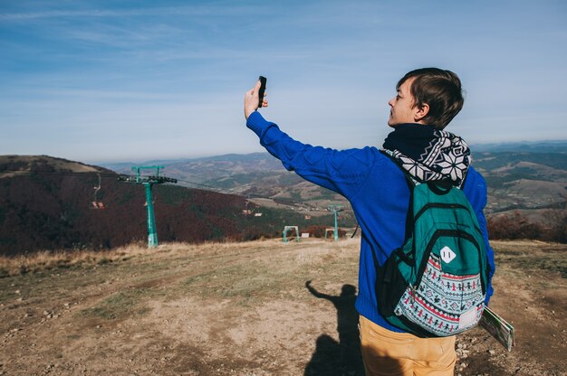 Young hipster taking photo by smart-phone on the peak of mountain