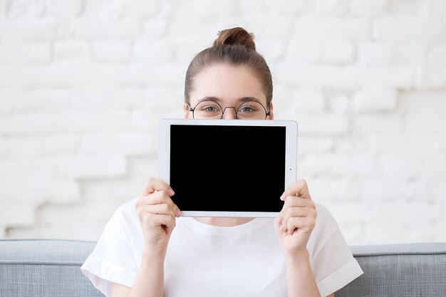 Foto un giovane adolescente hipster con un panino in occhiali rotondi e una maglietta bianca si nasconde dietro un tablet digitale con schermo nero per lo spazio di copia