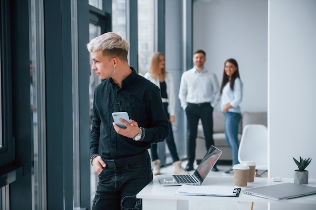 Young hipster with phone standing in front of group of young successful team that working and communicating together indoors in office