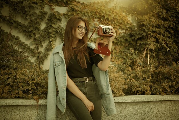 デニムジャケットとメガネの若い流行に敏感な女性は、屋外でフィルムレトロカメラを楽しんでいます
