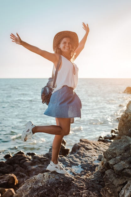 A young hipster woman in a hat and a rukzak with her hands up, standing on top of a cliff and looking at the sea at sunset.