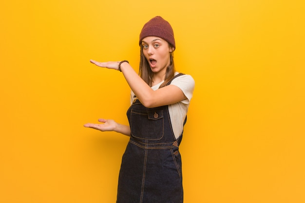 Young hipster woman holding something very surprised and shocked