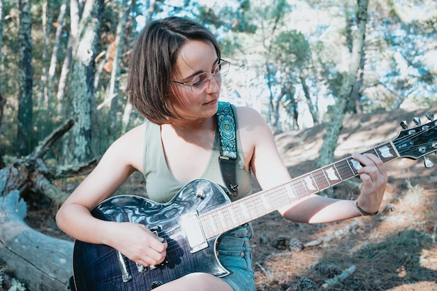 Young hipster woman playing the guitar outside the forest park of the city. Having fun learning a new skill, music play seasonal style. Young short hair girl. Copy space