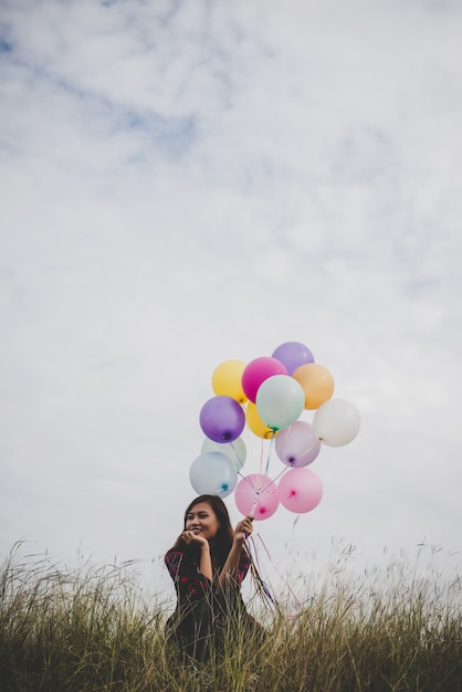 Young hipster woman sitting holding colorful balloons in field, blue sky background.