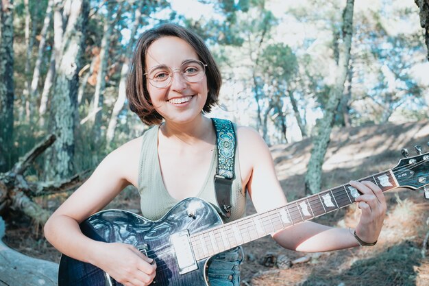 Young hipster woman sitting in a trunk playing guitar on park or garden background. Teen girl learning to play song and writing music. Hobby, lifestyle, relax, Instrument, leisure, education concept