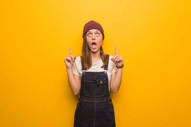 Young hipster woman surprised pointing up to show something