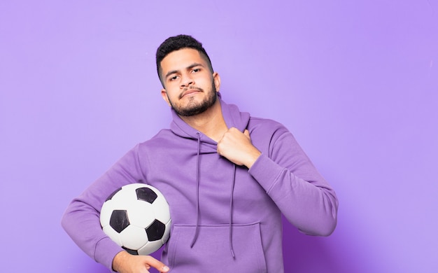 Young hispanic athlete man scared expression and holding a soccer ball