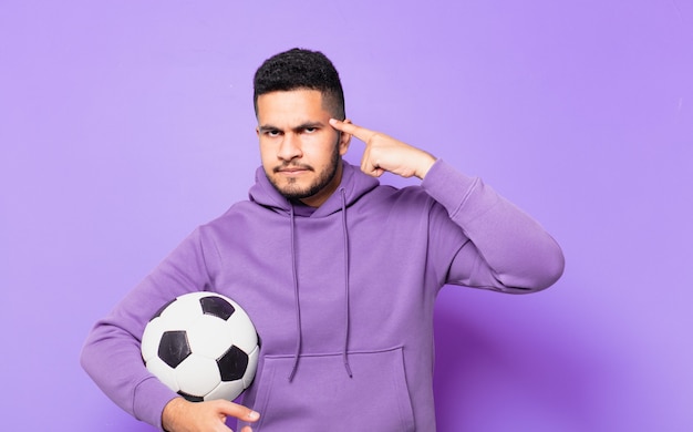 Young hispanic athlete man with thinking expression and holding a soccer ball