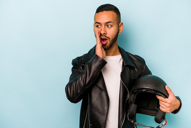 Young hispanic biker man isolated on blue background is saying a secret hot braking news and looking aside