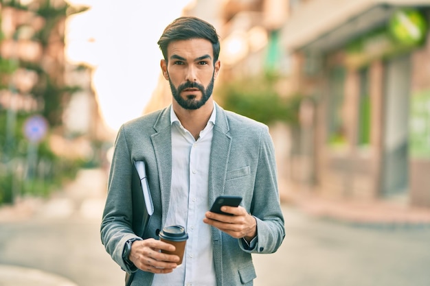 Молодой латиноамериканский бизнесмен с серьезным выражением лица пользуется смартфоном и пьет кофе в городе