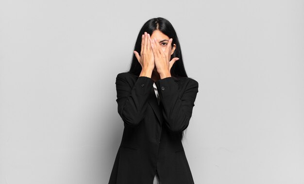 Young hispanic businesswoman covering face with hands, peeking between fingers with surprised expression and looking to the side