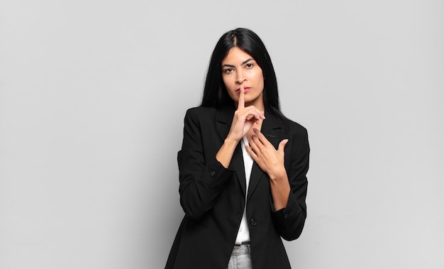 Young hispanic businesswoman looking serious and cross with finger pressed to lips demanding silence or quiet, keeping a secret