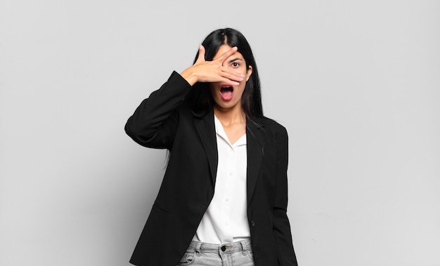 Young hispanic businesswoman looking shocked, scared or terrified, covering face with hand and peeking between fingers