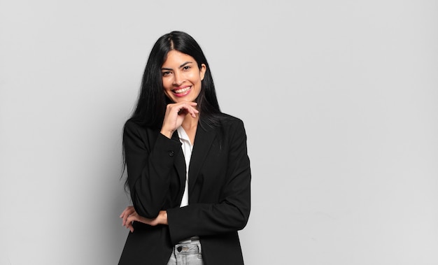 Young hispanic businesswoman smiling, enjoying life, feeling happy, friendly, satisfied and carefree with hand on chin