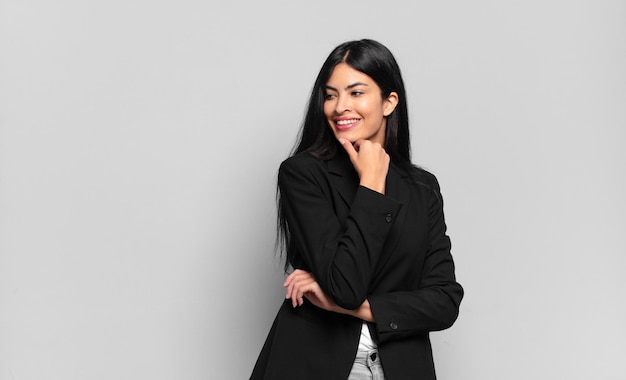 Young hispanic businesswoman smiling with a happy, confident expression with hand on chin, wondering and looking to the side