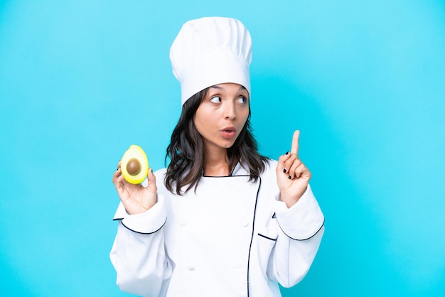 Photo young hispanic chef woman holding avocado isolated on blue background thinking an idea pointing the finger up