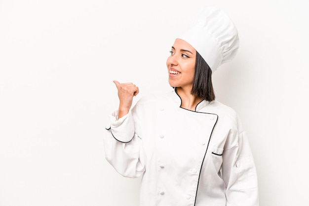 Young hispanic chef woman isolated on white background points with thumb finger away laughing and carefree