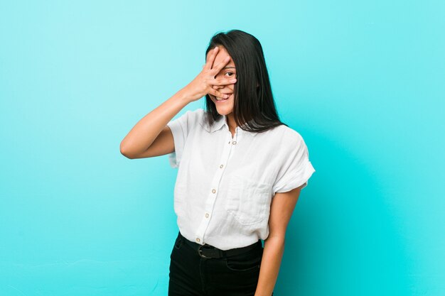 Young hispanic cool woman against a blue wall blink at the camera through fingers, embarrassed covering face.