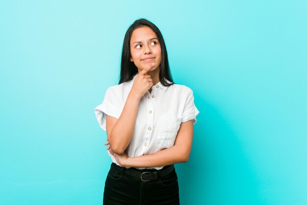 Young hispanic cool woman against a blue wall looking sideways with doubtful and skeptical expression.