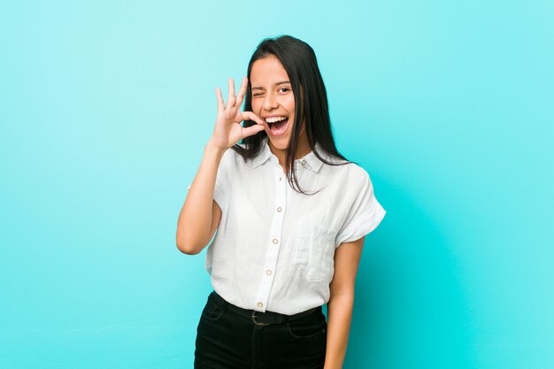 Young hispanic cool woman against a blue wall winks an eye and holds an okay gesture with hand.