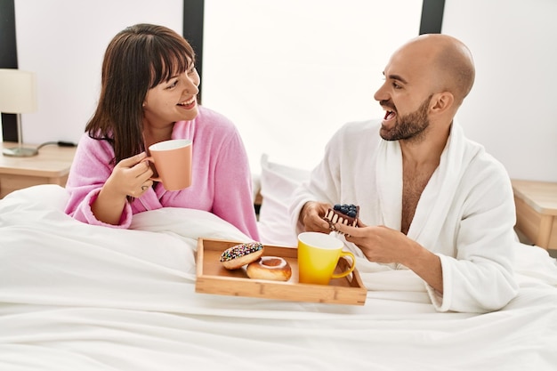 Young hispanic couple having breakfast sitting on the bed at bedroom.