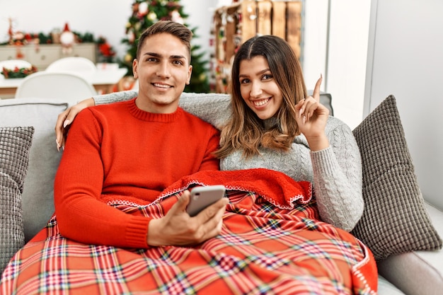 Молодая латиноамериканская пара, сидящая на диване на рождество, удивлена идеей или вопросом, указывая пальцем со счастливым лицом номер один