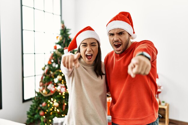 Young hispanic couple standing by christmas tree pointing displeased and frustrated to the camera, angry and furious with you