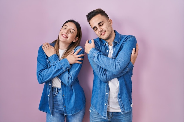 Young hispanic couple standing over pink background hugging oneself happy and positive, smiling confident. self love and self care