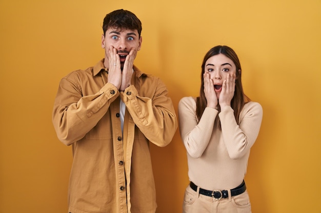 Young hispanic couple standing over yellow background afraid and shocked surprise and amazed expression with hands on face