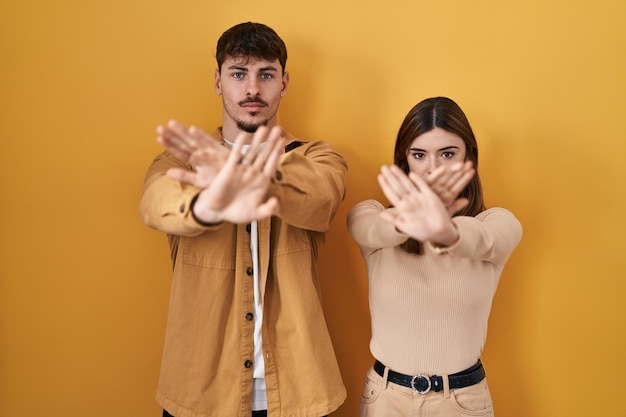 黄色の背景の上に立っている若いヒスパニック系カップルは、腕と手のひらを交差させ、否定的なサインをし、怒った顔をしている拒否表現