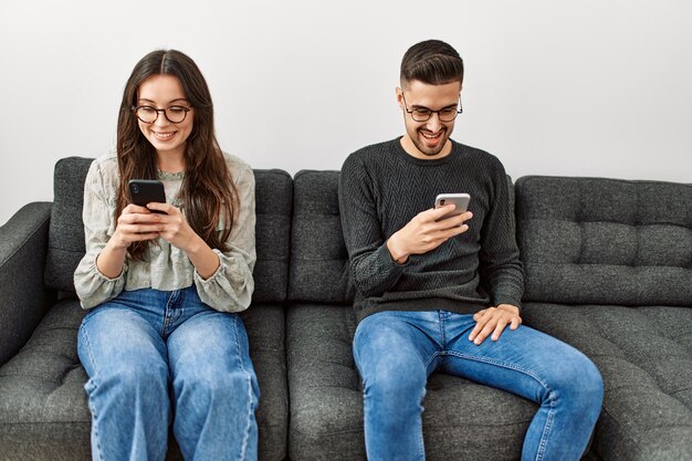 young hispanic couple using smartphone sitting on the sofa at home