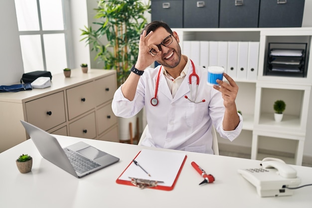 Young hispanic doctor man with beard holding ear cotton buds smiling happy doing ok sign with hand on eye looking through fingers