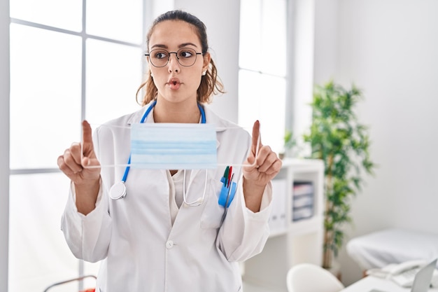 Young hispanic doctor woman holding safety mask making fish face with mouth and squinting eyes crazy and comical