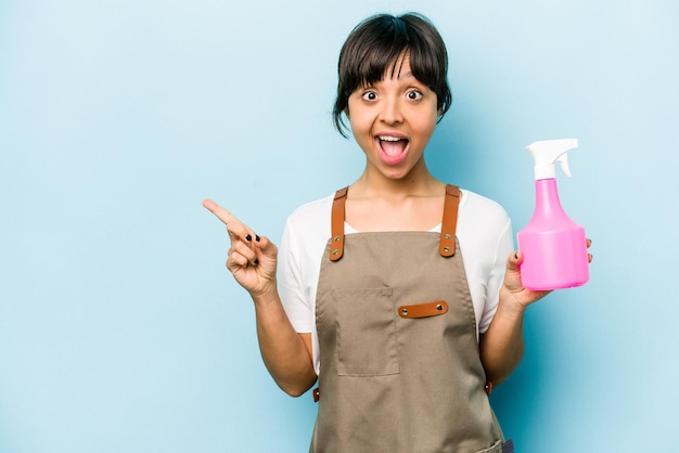 Young hispanic gardener woman holding a hand sprayer isolated on blue background pointing to the side