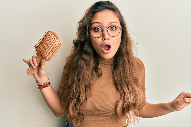 Young hispanic girl holding hair comb scared and amazed with open mouth for surprise, disbelief face