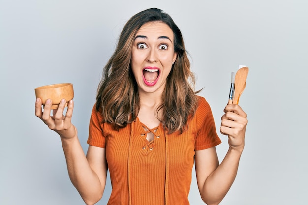 Young hispanic girl holding make up products celebrating crazy and amazed for success with open eyes screaming excited.