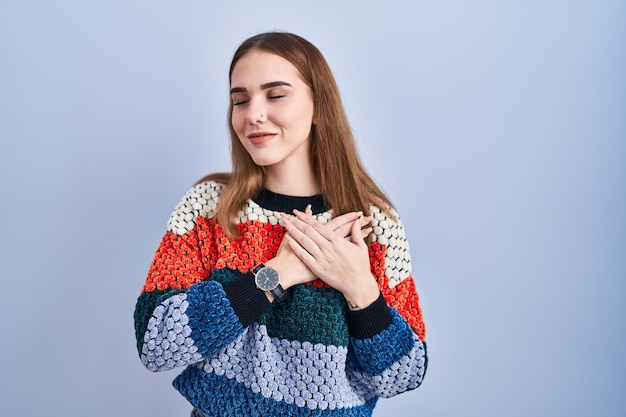 Young hispanic girl standing over blue background smiling with hands on chest with closed eyes and grateful gesture on face health concept