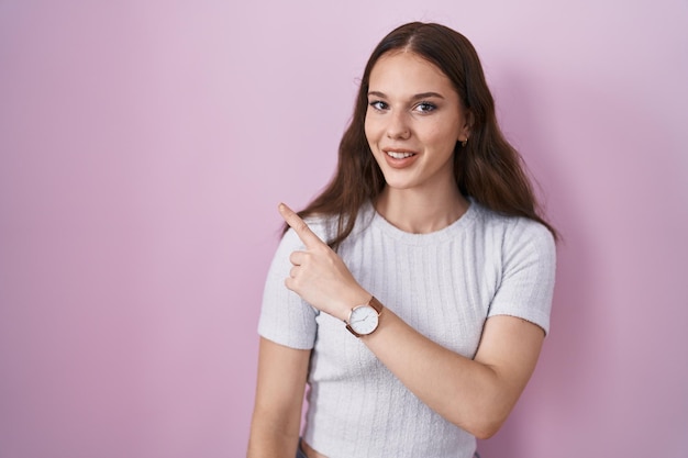 Photo young hispanic girl standing over pink background cheerful with a smile on face pointing with hand and finger up to the side with happy and natural expression