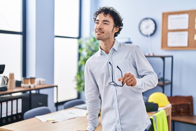 オフィスで眼鏡を持って自信を持って微笑む若いヒスパニック系男性建築家