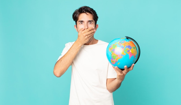 Young hispanic man covering mouth with hands with a shocked and holding a world globe map