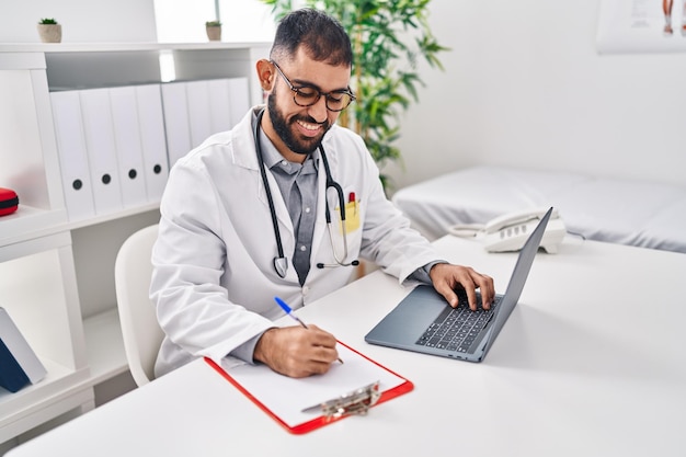Young hispanic man doctor using laptop writing medical report at clinic