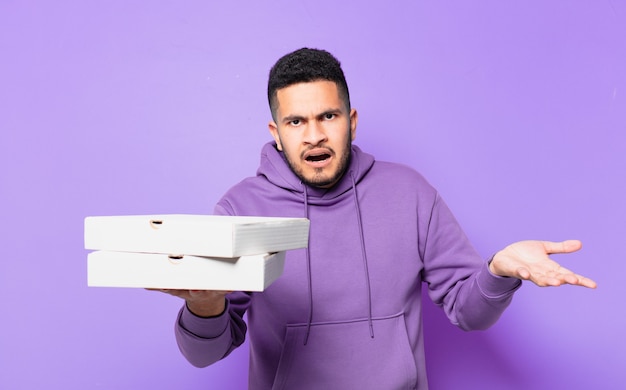 Young hispanic man doubting or uncertain expression and holding take away pizzas