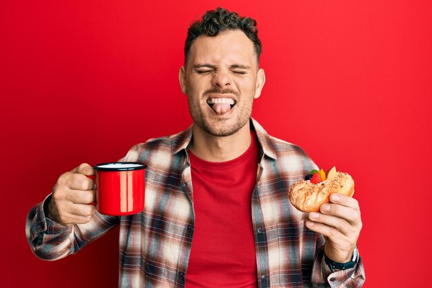 Photo young hispanic man drinking coffee and eating pastry sticking tongue out happy with funny expression