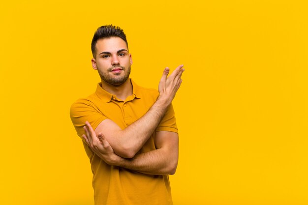 Young hispanic man feeling confused and clueless, wondering about a doubtful explanation or thought against orange wall