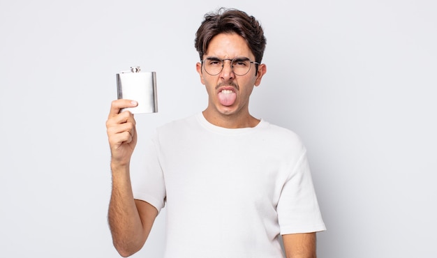 Young hispanic man feeling disgusted and irritated and tongue out. alcohol flask concept