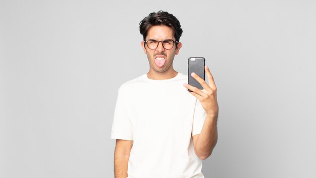Young hispanic man feeling disgusted and irritated and tongue out and holding a smartphone