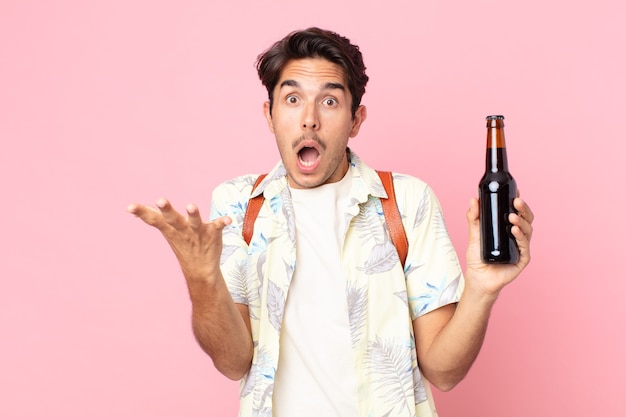 Young hispanic man feeling extremely shocked and surprised and holding a bottle of beer