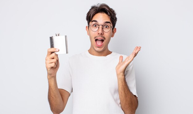 Young hispanic man feeling happy and astonished at something unbelievable. alcohol flask concept