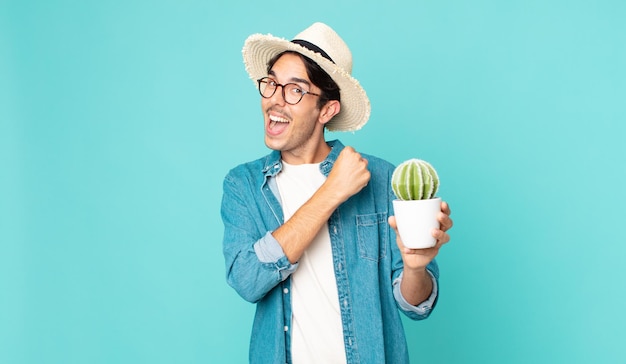 Young hispanic man feeling happy and facing a challenge or celebrating and holding a cactus